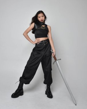 Full length portrait of pretty young asian girl wearing black tank top, utilitarian  pants and leather boots. Standing pose  holding a sword, isolated against a  studio background.