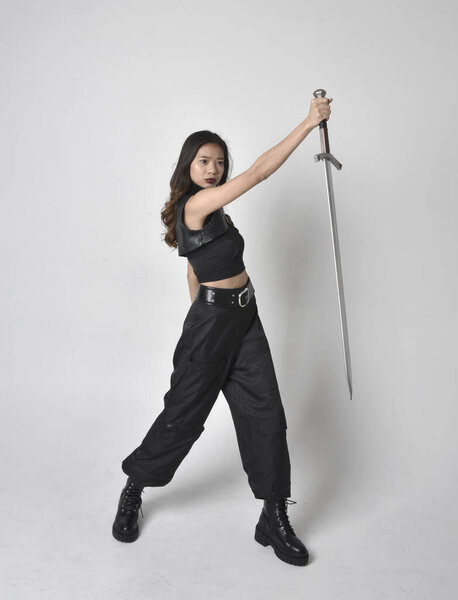 полный портрет симпатичной молодой азиатской девушки в черной майке, утилитарных брюках и кожаных сапогах. Стоя поза держа меч, изолированные на фоне студии.