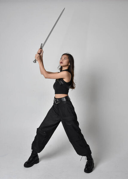 полный портрет симпатичной молодой азиатской девушки в черной майке, утилитарных брюках и кожаных сапогах. Стоя поза держа меч, изолированные на фоне студии.