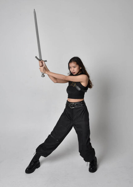полный портрет симпатичной молодой азиатской девушки в черной майке, утилитарных брюках и кожаных сапогах. Стоя поза держа меч, изолированные на фоне студии.