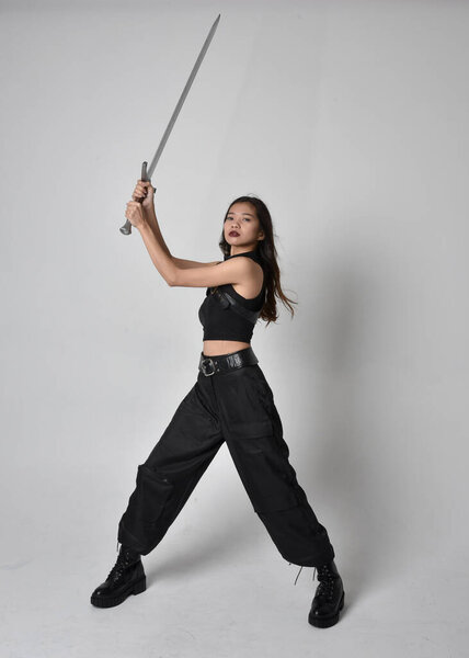 полный портрет симпатичной молодой азиатской девушки в черной майке, утилитарных брюках и кожаных сапогах. Стоя поза держа меч, изолированные на фоне студии.