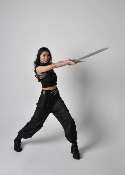полный портрет симпатичной молодой азиатской девушки в черной майке, утилитарных брюках и кожаных сапогах. Стоя поза держа меч, изолированные на фоне студии.