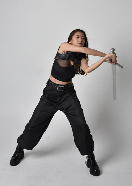 полный портрет симпатичной молодой азиатской девушки в черной майке, утилитарных брюках и кожаных сапогах. Стоя поза держа меч, изолированные на фоне студии.