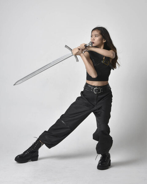 полный портрет симпатичной молодой азиатской девушки в черной майке, утилитарных брюках и кожаных сапогах. Стоя поза держа меч, изолированные на фоне студии.