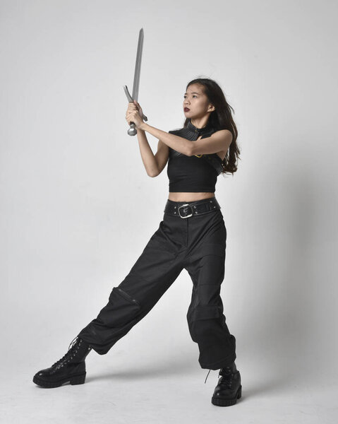 полный портрет симпатичной молодой азиатской девушки в черной майке, утилитарных брюках и кожаных сапогах. Стоя поза держа меч, изолированные на фоне студии.