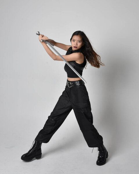 полный портрет симпатичной молодой азиатской девушки в черной майке, утилитарных брюках и кожаных сапогах. Стоя поза держа меч, изолированные на фоне студии.