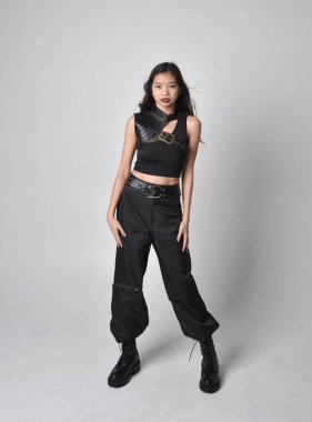 Full length portrait of pretty young asian girl wearing black tank top, utilitarian  pants and leather boots. Standing pose with gestural hands, isolated against a  studio background.