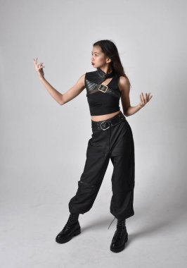 Full length portrait of pretty young asian girl wearing black tank top, utilitarian  pants and leather boots. Standing pose with gestural hands, isolated against a  studio background.