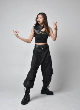 Full length portrait of pretty young asian girl wearing black tank top, utilitarian  pants and leather boots. Standing pose with gestural hands, isolated against a  studio background.