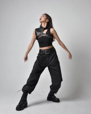 Full length portrait of pretty young asian girl wearing black tank top, utilitarian  pants and leather boots. Standing pose with gestural hands, isolated against a  studio background.