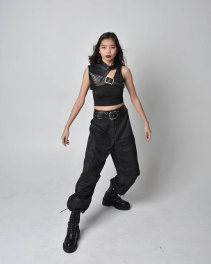 Full length portrait of pretty young asian girl wearing black tank top, utilitarian  pants and leather boots. Standing pose with gestural hands, isolated against a  studio background.