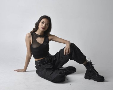 Full length portrait of pretty young asian girl wearing black tank top, utilitarian  pants and leather boots. Sitting poseisolated against a  studio background.
