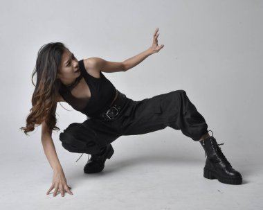 Full length portrait of pretty young asian girl wearing black tank top, utilitarian  pants and leather boots. Sitting poseisolated against a  studio background.