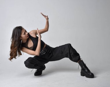 Full length portrait of pretty young asian girl wearing black tank top, utilitarian  pants and leather boots. Sitting poseisolated against a  studio background.