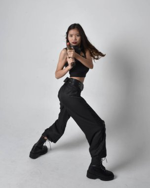 Full length portrait of pretty young asian girl wearing black tank top, utilitarian  pants and leather boots. Standing pose holding a gun,  isolated against a  studio background.