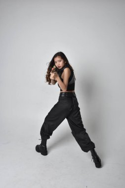 Full length portrait of pretty young asian girl wearing black tank top, utilitarian  pants and leather boots. Standing pose holding a gun,  isolated against a  studio background.