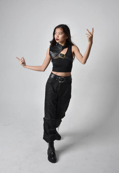 полный портрет симпатичной молодой азиатской девушки в черной майке, утилитарных брюках и кожаных сапогах. Стоя поза с жестом руки, изолированные на фоне студии.