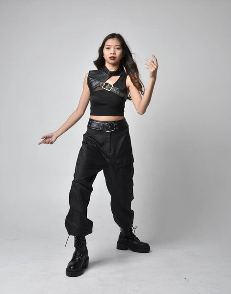 Full length portrait of pretty young asian girl wearing black tank top, utilitarian  pants and leather boots. Standing pose with gestural hands, isolated against a  studio background.