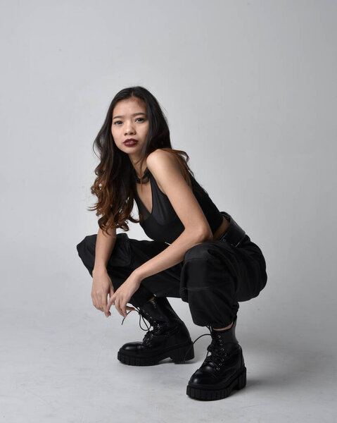 полный портрет симпатичной молодой азиатской девушки в черной майке, утилитарных брюках и кожаных сапогах. сидя на студийном фоне.