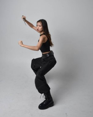 Full length portrait of pretty young asian girl wearing black tank top, utilitarian  pants and leather boots. Standing pose holding a knife,  isolated against a  studio background.