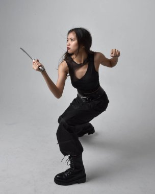 Full length portrait of pretty young asian girl wearing black tank top, utilitarian  pants and leather boots. Standing pose holding a knife,  isolated against a  studio background.