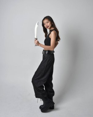Full length portrait of pretty young asian girl wearing black tank top, utilitarian  pants and leather boots. Standing pose holding a knife,  isolated against a  studio background.