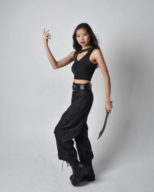 Full length portrait of pretty young asian girl wearing black tank top, utilitarian  pants and leather boots. Standing pose holding a knife,  isolated against a  studio background.