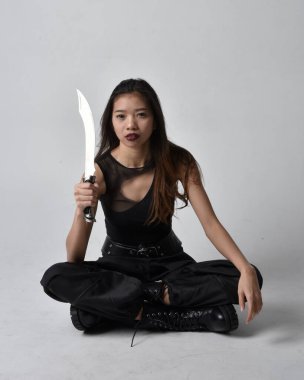 Full length portrait of pretty young asian girl wearing black tank top, utilitarian  pants and leather boots. Sitting pose holding a knife,  isolated against a  studio background.