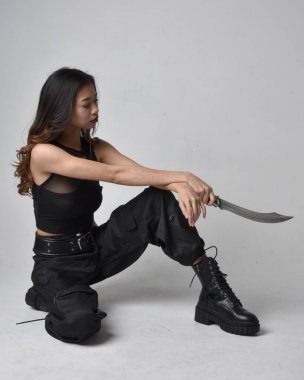Full length portrait of pretty young asian girl wearing black tank top, utilitarian  pants and leather boots. Sitting pose holding a knife,  isolated against a  studio background.