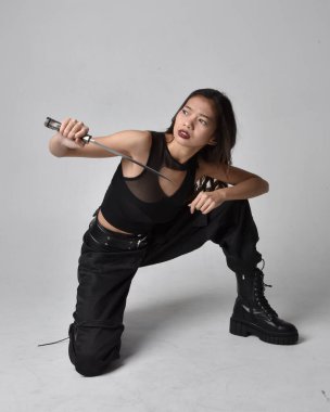 Full length portrait of pretty young asian girl wearing black tank top, utilitarian  pants and leather boots. Sitting pose holding a knife,  isolated against a  studio background.