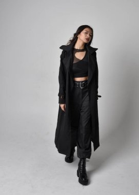 Full length portrait of pretty young asian girl wearing black tank top, utilitarian  pants, leather boots and large dark coat. Standing pose with gestural hand movements,  isolated on  studio background.