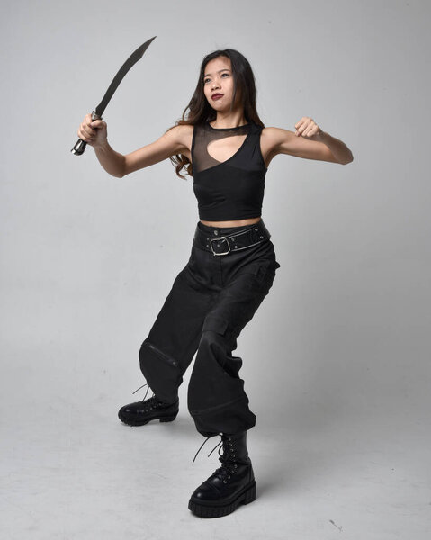 полный портрет симпатичной молодой азиатской девушки в черной майке, утилитарных брюках и кожаных сапогах. Стоя поза держа нож, изолированные на фоне студии.