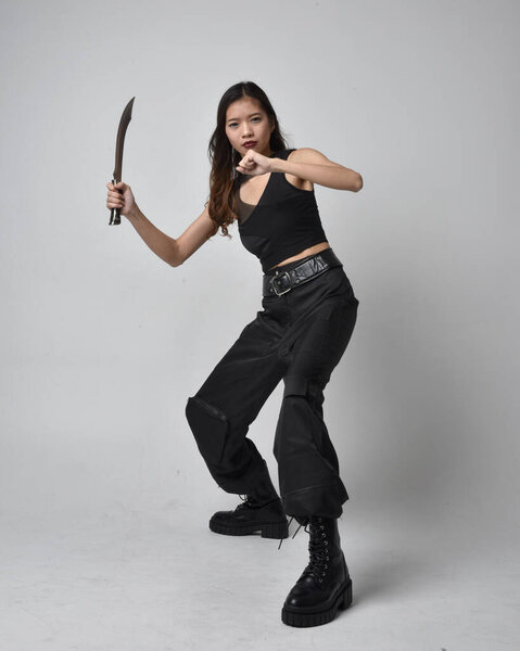 полный портрет симпатичной молодой азиатской девушки в черной майке, утилитарных брюках и кожаных сапогах. Стоя поза держа нож, изолированные на фоне студии.