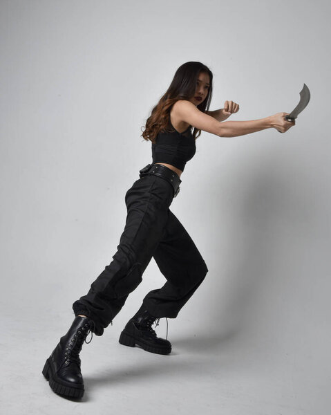 полный портрет симпатичной молодой азиатской девушки в черной майке, утилитарных брюках и кожаных сапогах. Стоя поза держа нож, изолированные на фоне студии.