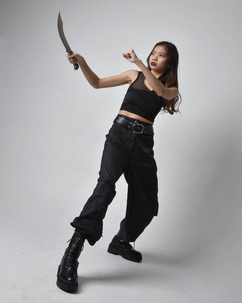 полный портрет симпатичной молодой азиатской девушки в черной майке, утилитарных брюках и кожаных сапогах. Стоя поза держа нож, изолированные на фоне студии.