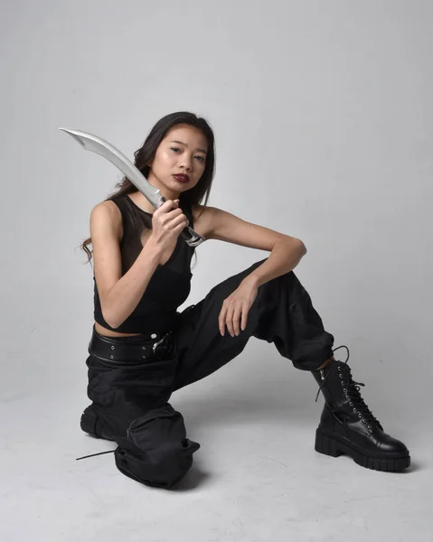 全长肖像漂亮的年轻亚洲女孩穿着黑色的坦克顶部 实用的裤子和皮靴 拿着刀坐着摆姿势 与工作室背景隔离 — 图库照片