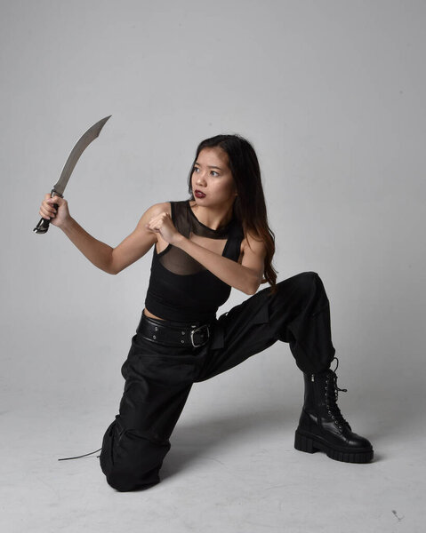 полный портрет симпатичной молодой азиатской девушки в черной майке, утилитарных брюках и кожаных сапогах. сидящая поза с ножом, изолированная от студийного фона.