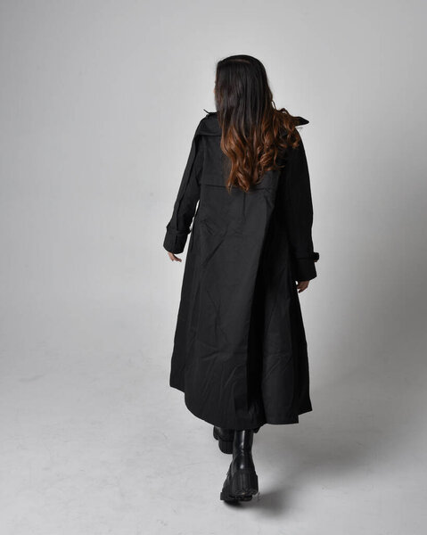 Полнометражный портрет симпатичной молодой азиатки в черной майке, утилитарных брюках, кожаных сапогах и большом темном пальто. Стоячая поза с жестом движения рукой, изолированные на фоне студии.