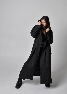 Full length portrait of pretty young asian girl wearing black tank top, utilitarian  pants, leather boots and large dark coat. Standing pose with gestural hand movements,  isolated on  studio background.
