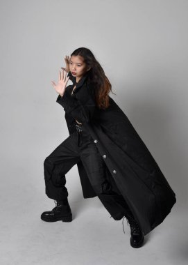 Full length portrait of pretty young asian girl wearing black tank top, utilitarian  pants, leather boots and large dark coat. Standing pose with gestural hand movements,  isolated on  studio background.