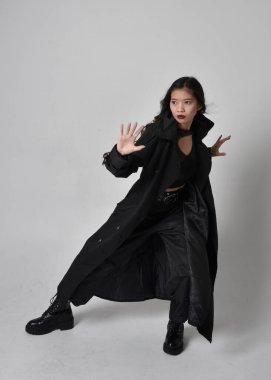 Full length portrait of pretty young asian girl wearing black tank top, utilitarian  pants, leather boots and large dark coat. Standing pose with gestural hand movements,  isolated on  studio background.
