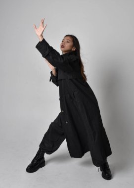 Full length portrait of pretty young asian girl wearing black tank top, utilitarian  pants, leather boots and large dark coat. Standing pose with gestural hand movements,  isolated on  studio background.