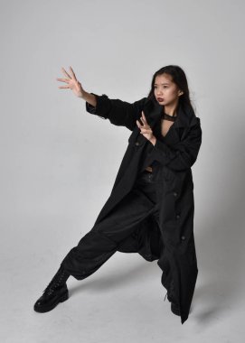 Full length portrait of pretty young asian girl wearing black tank top, utilitarian  pants, leather boots and large dark coat. Standing pose with gestural hand movements,  isolated on  studio background.
