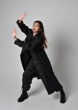 Full length portrait of pretty young asian girl wearing black tank top, utilitarian  pants, leather boots and large dark coat. Standing pose with gestural hand movements,  isolated on  studio background.