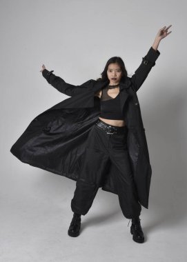 Full length portrait of pretty young asian girl wearing black tank top, utilitarian  pants, leather boots and large dark coat. Standing pose with gestural hand movements,  isolated on  studio background.
