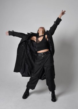 Full length portrait of pretty young asian girl wearing black tank top, utilitarian  pants, leather boots and large dark coat. Standing pose with gestural hand movements,  isolated on  studio background.