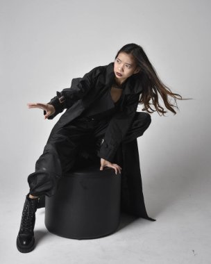 Full length portrait of pretty young asian girl wearing black tank top, utilitarian  pants, leather boots and large dark coat. Sitting pose with gestural hand movements,  isolated on  studio background.