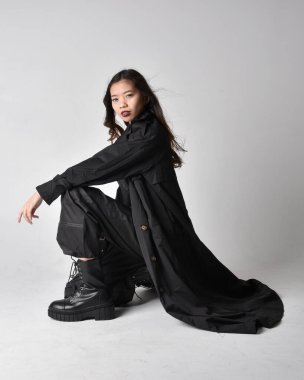 Full length portrait of pretty young asian girl wearing black tank top, utilitarian  pants, leather boots and large dark coat. Sitting pose with gestural hand movements,  isolated on  studio background.