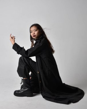 Full length portrait of pretty young asian girl wearing black tank top, utilitarian  pants, leather boots and large dark coat. Sitting pose with gestural hand movements,  isolated on  studio background.
