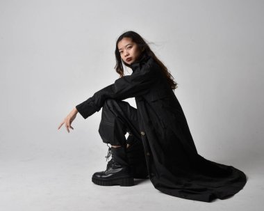 Full length portrait of pretty young asian girl wearing black tank top, utilitarian  pants, leather boots and large dark coat. Sitting pose with gestural hand movements,  isolated on  studio background.